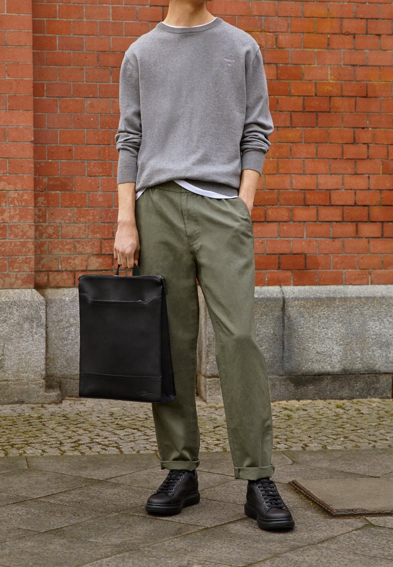 Grijze katoenen trui, olijfgroene broek en zwarte hoge sneakers. Zwarte leren tas met een voorkantzak, tegen een bakstenen muur.