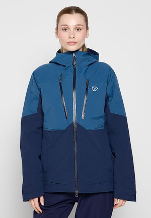 Frau trägt eine zweifarbige blaue Kapuzenjacke mit vorderen Reißverschlusstaschen und einem kleinen Logo auf der Brust, steht vor einem schlichten grauen Hintergrund.