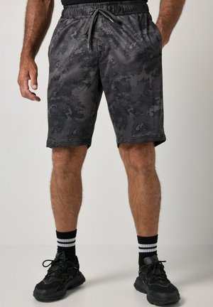 Mann trägt schwarze und graue Camouflage-Sportshorts, schwarze Sportschuhe und schwarze Socken mit weißen Streifen und steht vor einem schlichten Hintergrund.