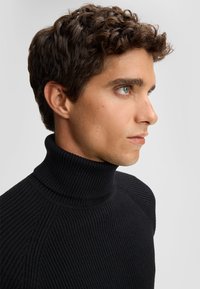 Maglione nero a collo alto con coste, caratterizzato da una vestibilità aderente e dettagli texture nel tessuto. Dettaglio ravvicinato del colletto.