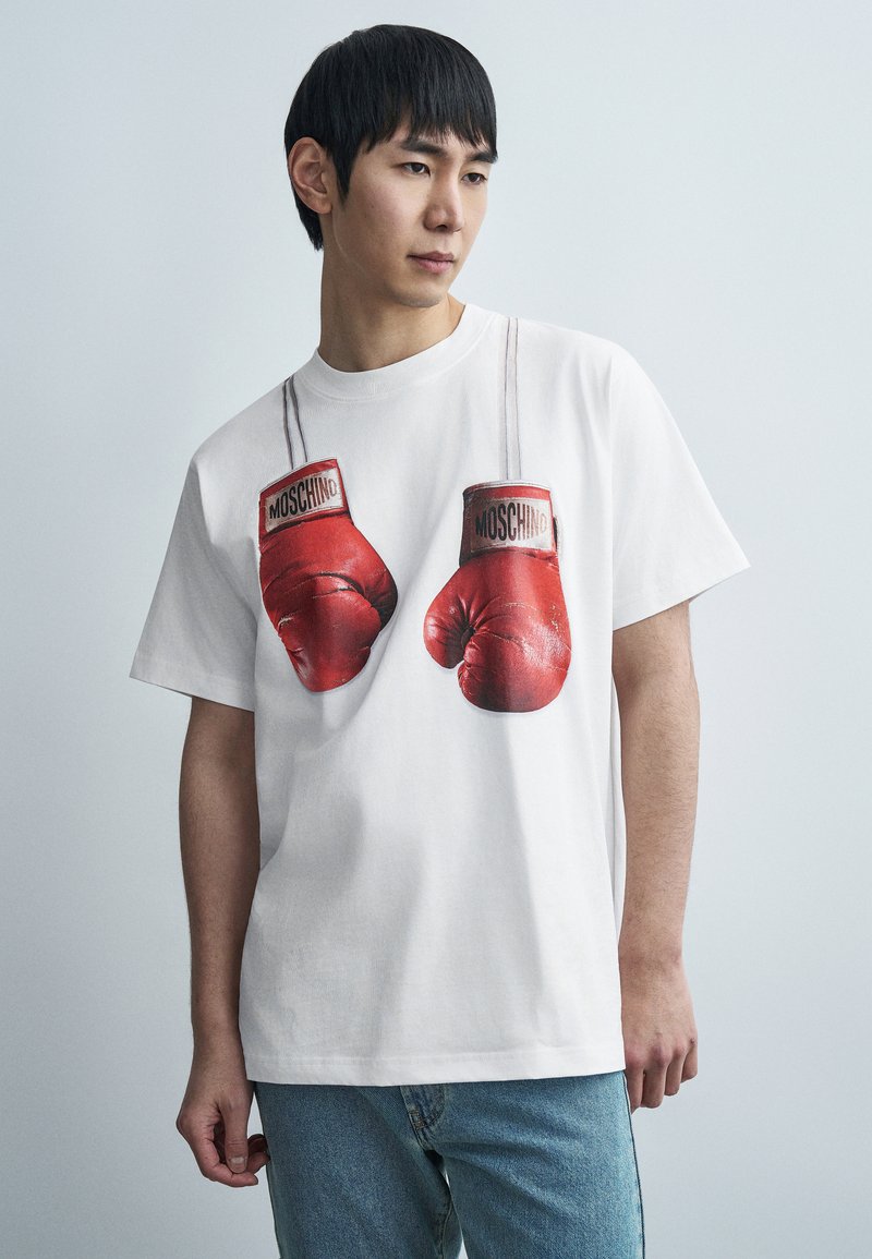 Λευκό βαμβακερό μπλουζάκι με γραφικό τύπωμα κόκκινων γαντιών πυγμαχίας και ετικέτα "MOSCHINO", στρογγυλή λαιμόκοψη, κοντά μανίκια και άνετη εφαρμογή.