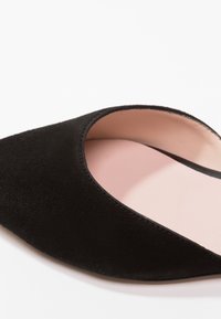 Chaussure plate en suédine noire avec un bout pointu et une doublure intérieure rose pâle sur fond blanc.