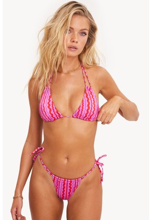 Femme blonde ajustant un bikini à motifs ondulés roses et rouges avec des liens sur les hanches, portant un bracelet en or et des boucles d'oreilles créoles sur fond blanc.