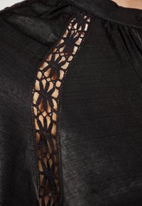 Schwarze Bluse mit strukturiertem Stoff und einem Spitzenbesatz an der Seite, der ein florales Muster und einen Kontrast zu dem glatten Material zeigt.