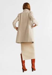 Manteau beige à col montant, détail poche au dos, associé à une jupe crème et des bottes en cuir marron montant jusqu'aux genoux. Texture lisse, coupe ajustée.