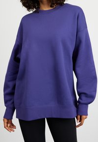 Sudadera de cuello redondo en color púrpura con mangas largas y sueltas, y un corte relajado. Confeccionada en un tejido suave con puños y dobladillo acanalados.