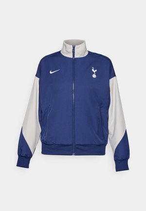 Chaqueta azul marino con cremallera y mangas grises, con un alto cuello, logo de Nike y emblema del Tottenham Hotspur. Fabricada con tejido suave.
