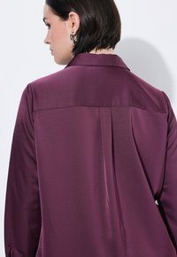 Pflaumenfarbene Satinbluse mit spitzem Kragen und Rückennahtdetails. Glatte Textur, lange Ärmel und Knopfmanschetten.