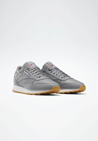 Pelēkas kedas ar gludu ādas augšdaļu, baltu vidusmēru un gumijas zoli. Iezīmes ietver apaļas kurpes diegus un logo detaļas uz mēles.