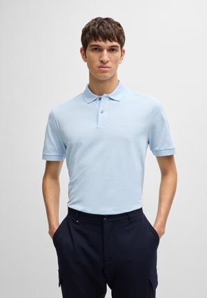 Polo de color azul claro hecho de una tela suave, con un cuello clásico, mangas cortas y un logo sutil en el pecho. Combinado con pantalones oscuros.