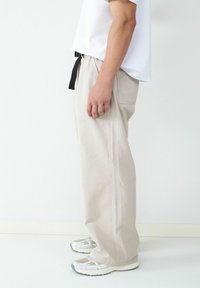 Persoon in beige baggy broek, witte sneakers en een wit shirt, die zijwaarts staat tegen een effen witte achtergrond.