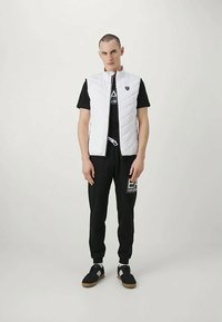 Vīrietis stāv pretī vienkāršai gaišai fona, ģērbies baltā quiltētā veste, melnā T-kreklā, melnās sporta biksēs ar logo un melnās kedās ar baltām akcentiem.