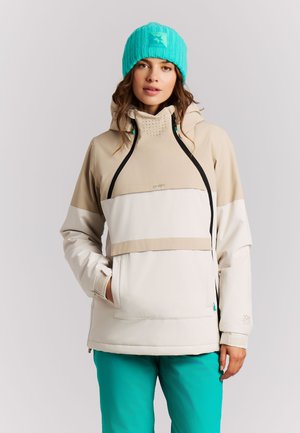 Vrouw die een beige-witte winterjas met zwarte ritsen draagt, petrolkleurige broek en een petrolkleurige gebreide muts, staand tegen een lichtgrijze achtergrond.