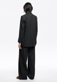 Traje de cuadros oscuros que incluye un blazer largo con abertura y pantalones de pierna ancha. El material parece texturizado con un diseño estructurado.