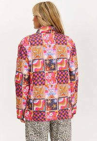 Kolorowa, wzorzysta kurtka z różnorodnymi kształtami geometrycznymi, kwiatami i abstrakcyjnymi wzorami, wykonana z gładkiego, lekkiego materiału.