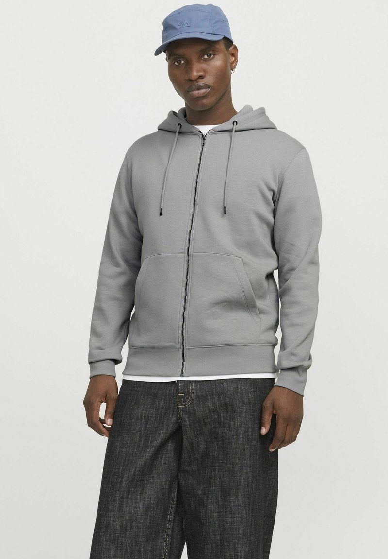Helles grau Zip-Hoodie mit einer verstellbaren Kapuze, vorderen Taschen und einer bequemen Passform, kombiniert mit dunklen Jeans und einer blauen Kappe.