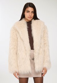 Cappotto in pelliccia sintetica color crema, con una texture shaggy, revers ampi e maniche lunghe. Indossato su un top marrone e una gonna beige.