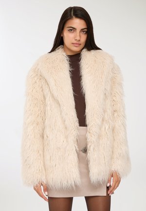 Cappotto in pelliccia sintetica color crema, con una texture shaggy, revers ampi e maniche lunghe. Indossato su un top marrone e una gonna beige.