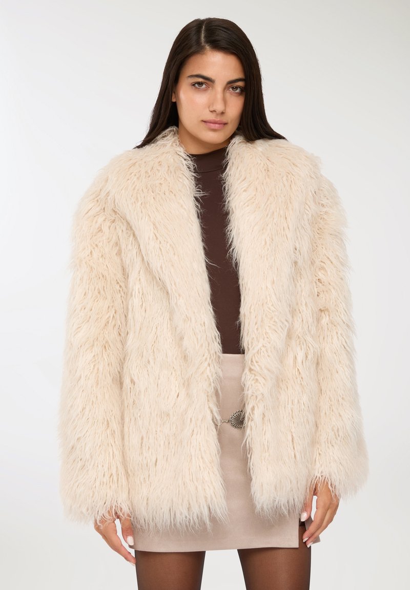 Cappotto in pelliccia sintetica color crema, con una texture shaggy, revers ampi e maniche lunghe. Indossato su un top marrone e una gonna beige.