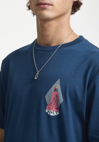 Blå bomulds T-shirt med et frontgrafik, der viser en pink figur og geometriske linjer. Rund hals og korte ærmer, glat tekstur.