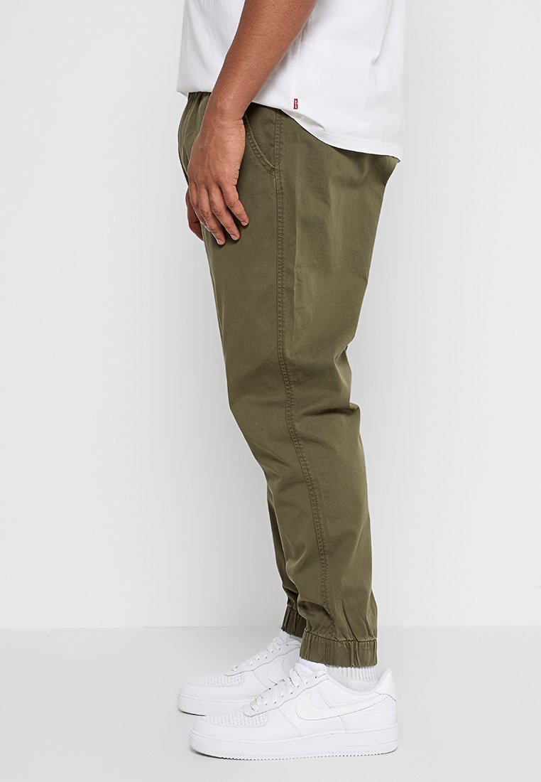 Pantaloni a gamba affusolata verde oliva con vita e polsini elasticizzati, realizzati in tessuto di cotone. Indossati con sneakers bianche per un look casual.