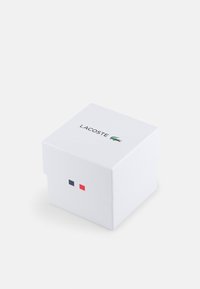 Caja cuadrada blanca con tapa abatible, que presenta el logotipo "LACOSTE" y un gráfico de cocodrilo verde; adornada con pequeños cuadrados rojos y azules.