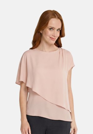Woman with long red hair wearing light pink asymmetrical short-sleeve blouse and dark pants, smiling against plain white background.