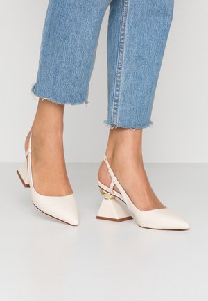 Stopy w beżowych sandałach na obcasie typu slingback z geometrycznym, blokowym obcasem, zestawione z niebieskimi, przyciętymi dżinsami z postrzępionym dołem na białej podłodze.