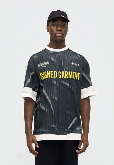 Νεαρός άνδρας που φοράει μια μαύρη και λευκή φαρδιά μπλούζα Moschino με κίτρινο κείμενο «SIGNED GARMENT» και ένα μαύρο καπέλο Moschino.