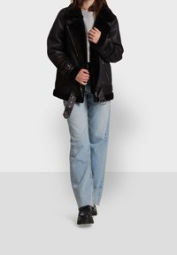 Svart shearlingjacka med bred krage, silverdragkedjor och justerbara remmar, tillsammans med ljusblå denimjeans och svarta stövlar.