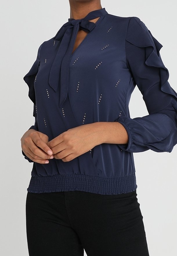 Femme portant un chemisier bleu marine à manches volantées, avec un nœud noué au col et un motif de petits points dispersés, associé à un pantalon noir.