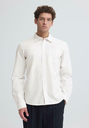 Junger Mann mit lockigem Haar, der ein cremefarbenes, vertikal gestreiftes Hemd mit Brusttasche und dunkle Hose vor einem schlichten Hintergrund trägt.