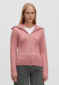 Frau, die einen rosa gerippten Reißverschluss-Pullover mit einem breiten Kragen, silbernen Creolen, einem weißen Oberteil und dunkelgrauen Jeans trägt, steht vor einem schlichten Hintergrund.