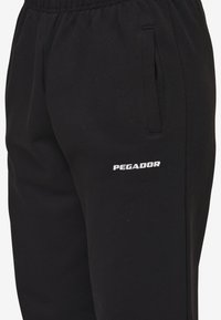 Zwarte joggingbroek gemaakt van zachte stof met een elastische tailleband, voorzien van zijzakken en een wit "PEGADOR" logo op de bovenbeen.