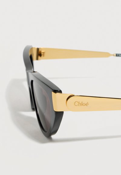 Μαύρα γυαλιά ηλίου με παχιά πλαίσια και χρυσά πλαϊνά μέρη που φέρουν το εγχάρακτο όνομα της μάρκας "Chloé" σε λευκό φόντο.