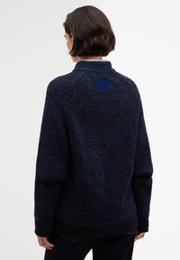 Marineblauwe gebreide trui met een gestructureerd patroon, voorzien van een hoge kraag, drop shoulders en een accenten vierkant detail op de achterkant.
