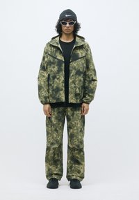 Camouflage-jack en -broek in groentinten, met een rits aan de voorkant, zwarte accenten, en gecombineerd met zwarte schoenen en een gebreide muts.