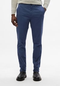 Schmal geschnittene blaue Chinos aus Baumwolle mit seitlichen Taschen und einem Knopfverschluss. Kombiniert mit schwarzen Schuhen, die eine glatte Textur und einen leichten Glanz aufweisen.
