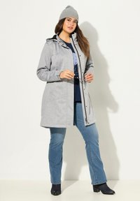 Chaqueta con capucha de color gris claro, con cremallera frontal, bolsillos laterales y detalles en negro. Combinada con una camisa de color azul marino y jeans azules. Gorro de punto gris.