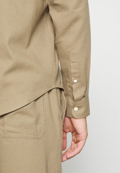 Camisa de manga longa marrom claro com textura suave, punhos dobráveis e dois botões. A costura lateral tem uma bainha arredondada, com detalhe de bolso visível.
