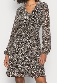 Robe à motifs noirs et beiges avec manches longues, décolleté en V et taille cintrée, en tissu fluide et léger.