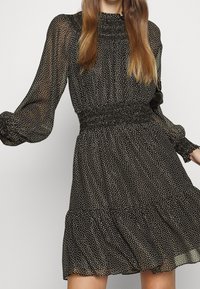 Femme portant une robe noire à manches longues avec des pois blancs, un ourlet à volants, une taille et des poignets smockés, debout devant un fond uni.