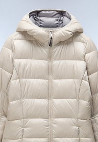 Chaqueta acolchada beige con capucha y diseño de acolchado. Presenta un cierre frontal, textura suave y cremalleras en los bolsillos laterales para funcionalidad.