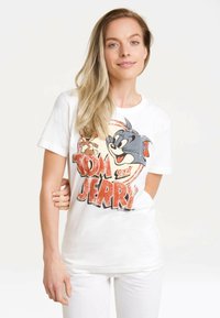 White cotton t-shirt featuring a large graphic print of cartoon characters Tom and Jerry, with bold orange and black lettering. Short sleeves.