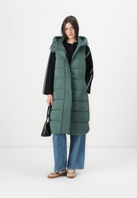 Grønn, polstret lang vest med hette, frontlukking med knapper, med et quiltet design. Brukt over en svart topp og kombinert med vide blå jeans.