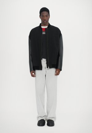 Black bomber jacket with leather sleeves, front zipper, and patch pocket over a gray sweater with red stripes, paired with light gray jeans.