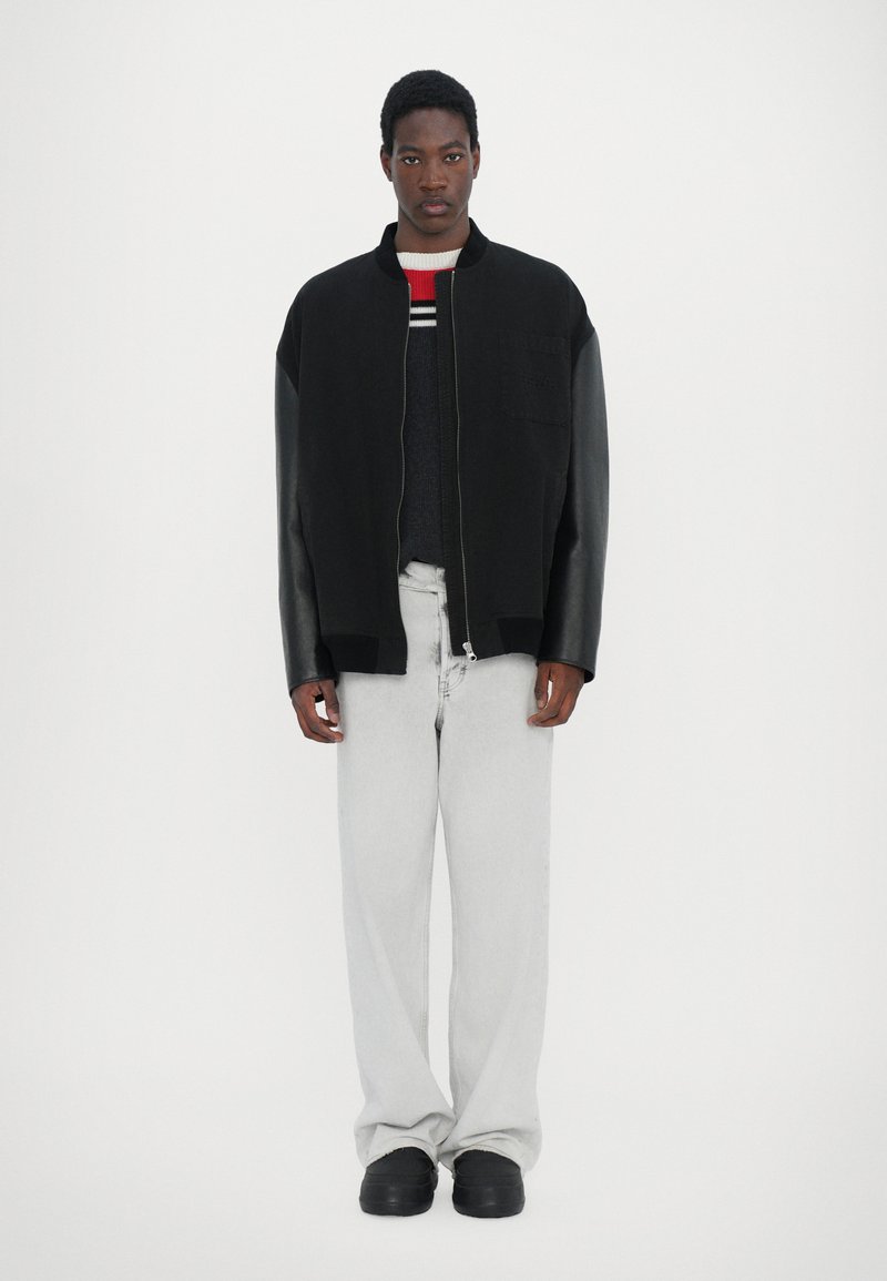 Black bomber jacket with leather sleeves, front zipper, and patch pocket over a gray sweater with red stripes, paired with light gray jeans.
