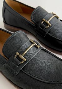 Mustad musta nahaga sussid, millel on tekstureeritud viimistlus. Rihmal on messingist pandla aksessuaar. Pruun välisolek, ümar varvas.