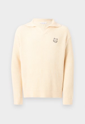 Mladý muž v krémovom pletenom svetri s logom líšky, široké hnedé nohavice s bočnými pruhmi, čierne topánky a modrý taška na ruky.