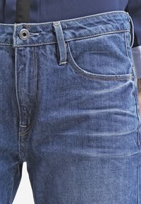 Vaqueros de mezclilla azul oscuro con una textura suave, que presentan un diseño clásico de cinco bolsillos y un cierre de botón plateado en la cintura.